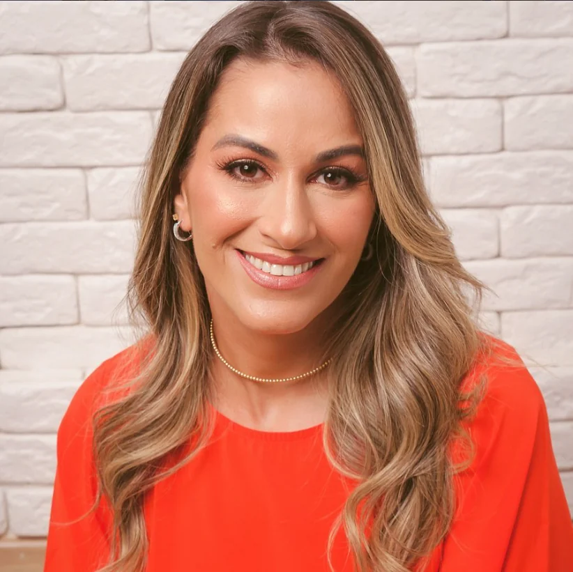 Aline Zia com blusa coral diante de parede clara